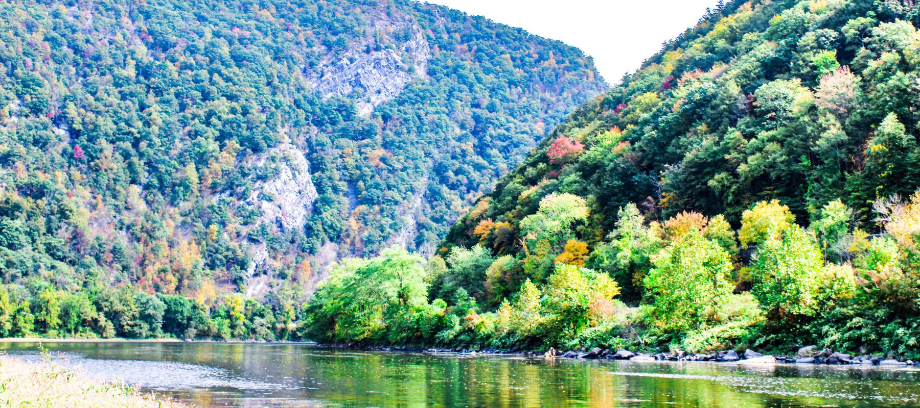 A picturesque river landscape flanked by steep, forested hills showing early signs of autumn. The foreground features a calm river reflecting the colorful foliage and the rugged, rocky hillside, creating a serene and natural setting