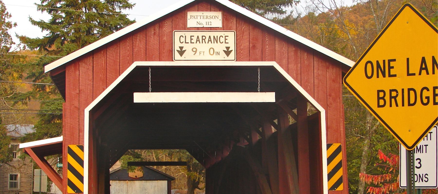 quaint red covered bridge, labeled as 'Pattison No. 112', with a clearance of 9 feet, located in a serene autumn setting. Adjacent to the bridge are warning signs including a 'One Lane Bridge' sign and a yellow weight limit sign indicating '3 Tons', nestled among vibrant fall foliage