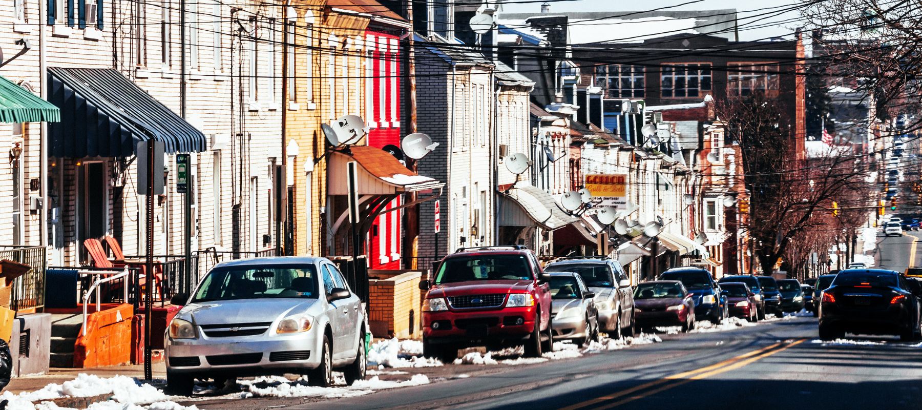 A vibrant urban street scene in winter, showcasing a row of colorful, tightly packed houses and businesses with various satellite dishes mounted on the facades. Cars are parked along the snow-lined street under a clear blue sky, reflecting the lively, residential character of the neighborhood