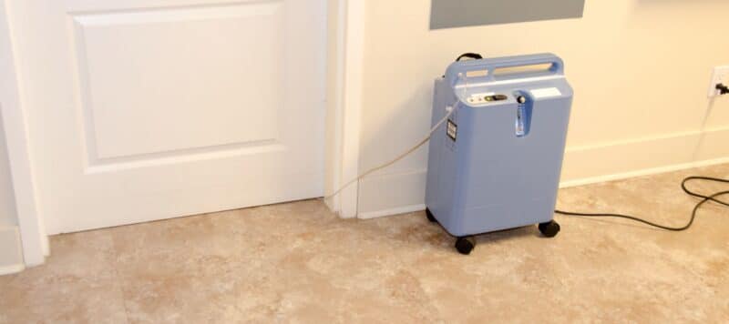 Portable healthcare oxygen machine in a room, plugged into a wall socket, standing on a beige carpet near a white door.