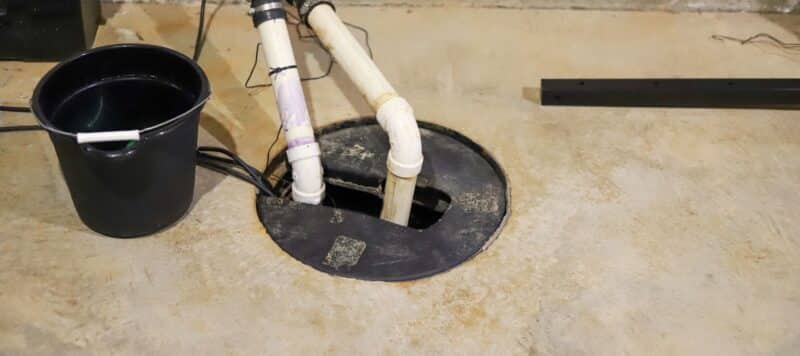 Sump pump installation in a basement with exposed pipes and electrical wiring, next to a black bucket on a concrete floor